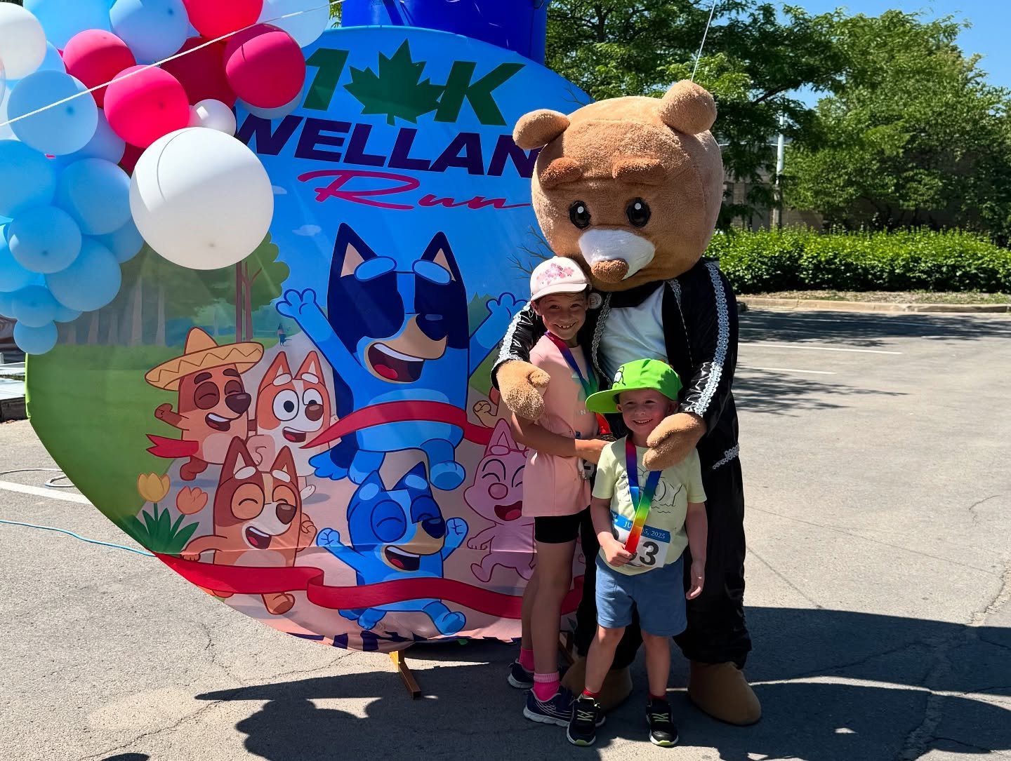 Two children pose with a teddy-bear mascot beside a colorful cartoon-dog event backdrop and balloons.