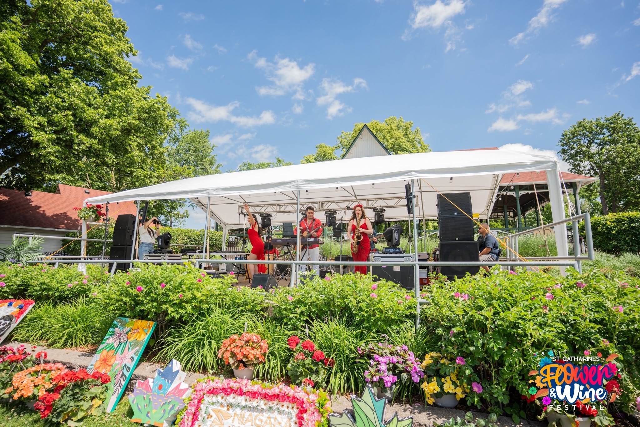 Outdoor flower festival stage with musicians performing amid colorful flower gardens and trees.