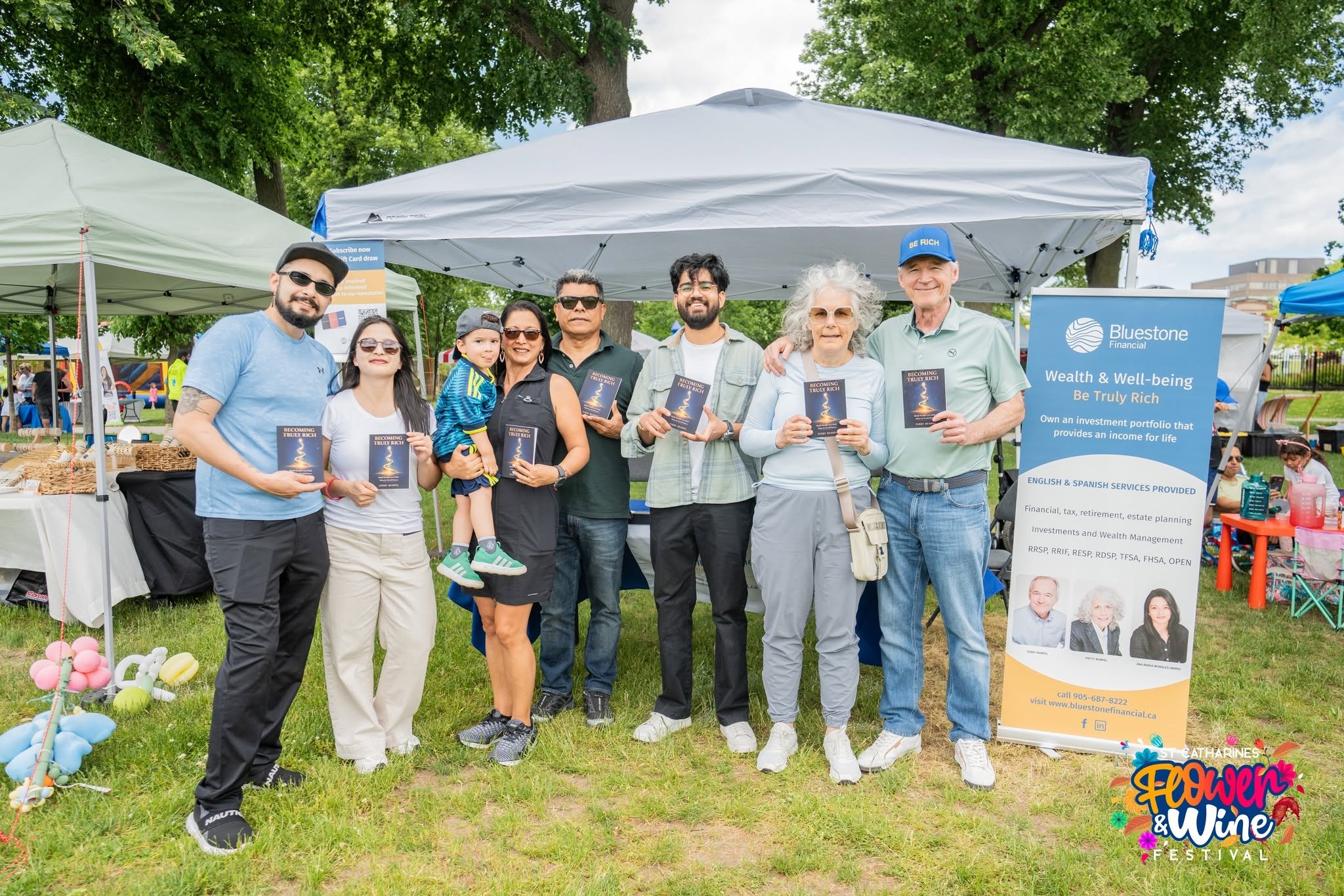 Eight people holding “BECOMING TRULY RICH” books at a festival booth beside a Bluestone Financial banner.
