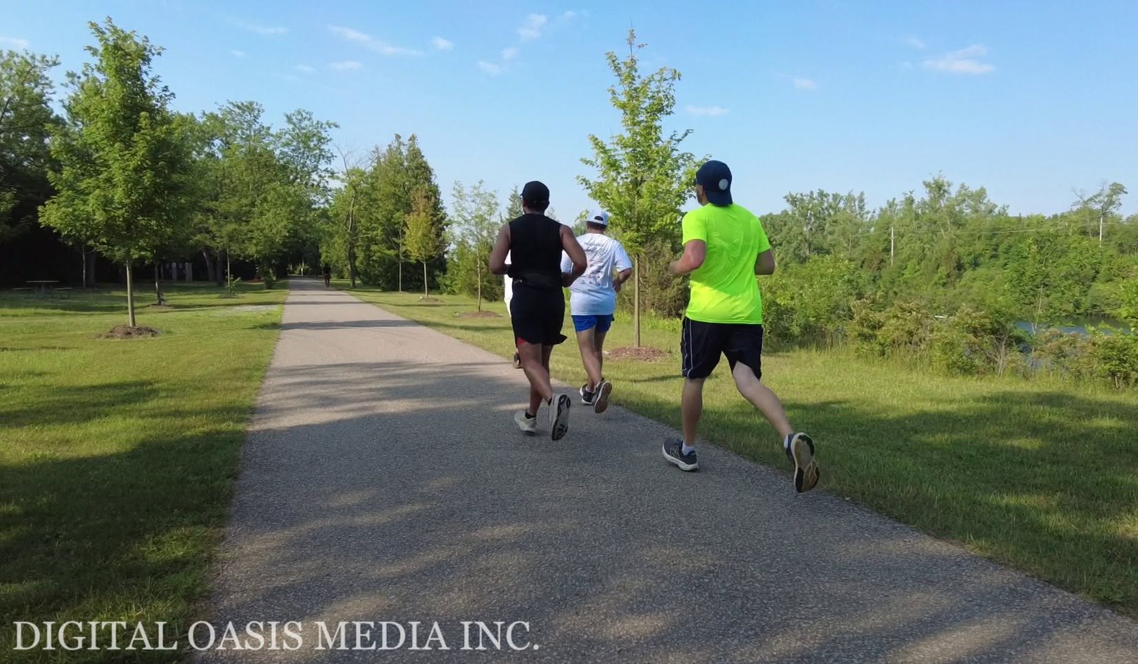 Three runners jog away on a paved park path bordered by trees, grass, and a partially visible pond.