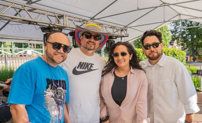 Four adults wearing sunglasses pose together under an outdoor event canopy.