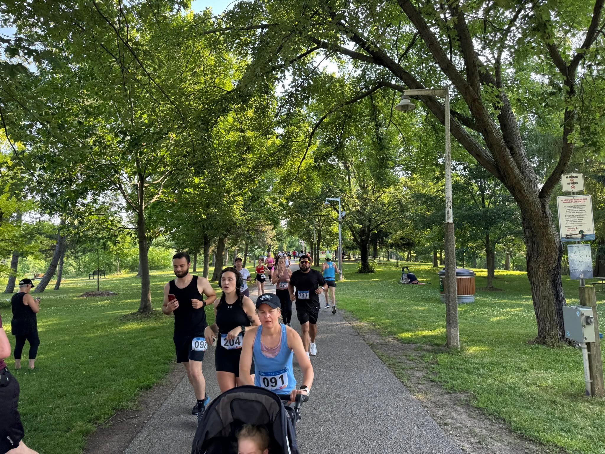 Runners move along a tree-lined park path, including a woman pushing a stroller with a race bib.